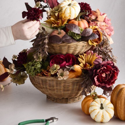 Hands arrange a lush autumn display in stacked wicker baskets, filled with pumpkins, dahlias, fall leaves, and deep burgundy blooms.
