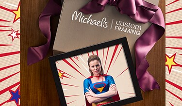 Michael’s Custom Framing ad featuring a framed superhero woman with folded arms, comic-book rays, Fan Expo Canada logo, wood backdrop and ribbons.