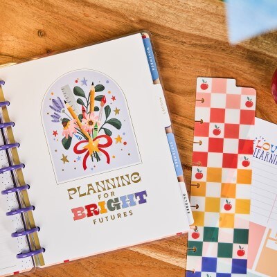 monthly planner with pencil crayon and ruler bouquet on wood table next to multicolored plaid apple divider