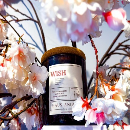 MakerPlace by Michaels A black candle labeled "WISH" with a cork lid is surrounded by blooming pink and white flowers. The setting is vibrant, fresh, and peaceful.