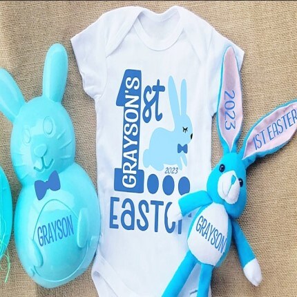 MakerPlace by Michaels White baby onesie with "Grayson's 1st Easter 2023" in blue, featuring a bunny. Beside it are a blue bunny toy and egg, both labeled "Grayson."
