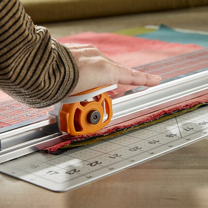 A person’s hand using an orange rotary cutter on a fabric laid over a cutting mat with measurement markings. The setting is bright and focused on crafting.
