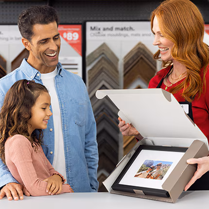 A Michaels employee showing a father and daughter a finished custom frame inside an open box.