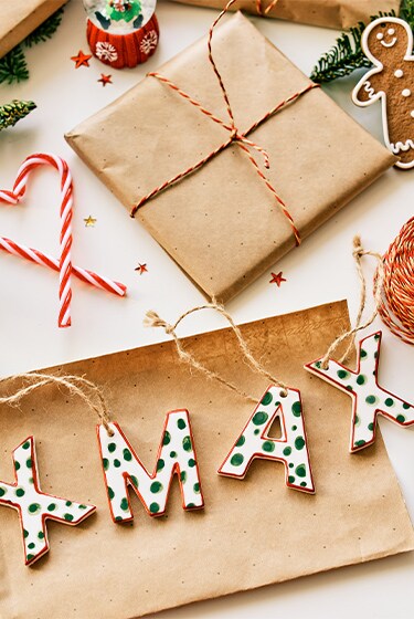 MakerPlace by Michaels Rustic Christmas gift-wrapping scene with twine, scissors, greenery, snowflake ornaments, and wooden “XMAS” letters, warm and festive.
