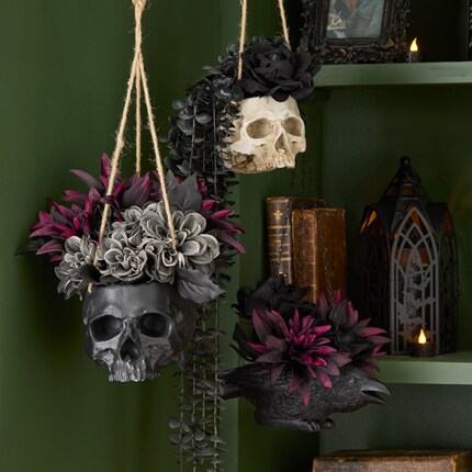 Halloween hanging planters featuring a black skull, a white skull, and a black crow, each adorned with dark purple and gray artificial flowers. 
