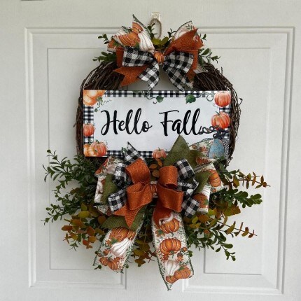 MakerPlace by Michaels Autumn wreath on a white door featuring a “Hello Fall” sign, orange pumpkins, plaid bows, ribbons, and lush greenery for a cozy welcome.