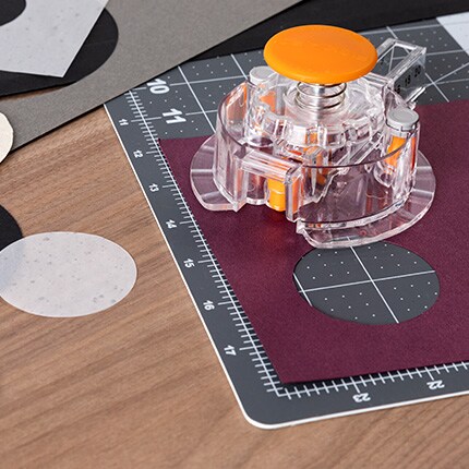 A clear circle cutter with an orange handle rests on a cutting mat, surrounded by cut paper circles. The scene is organized and creative.