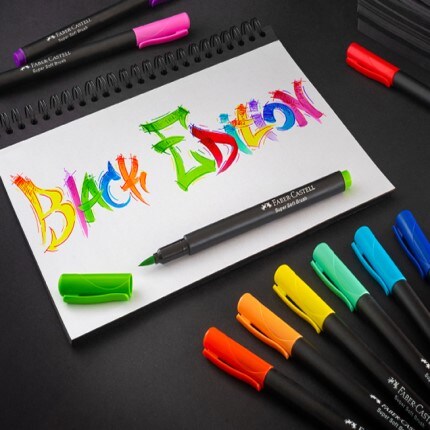 Black edition written on white paper with a black background with the Faber Castell Black Edition markers on the table around them