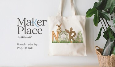 MakerPlace by Michaels A tote bag with the name "Nora" decorated with woodland animals stands against a light wall. Text reads "MakerPlace by Michaels, Handmade by: Pop Of Ink." A plant and woven basket are nearby.