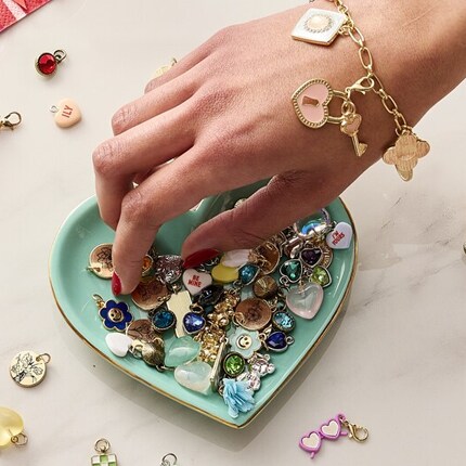 A hand wearing a charm bracelet reaches into a bowl of charms.