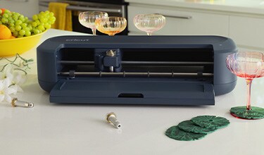 Open black Cricut cutting machine on a kitchen counter, with wine glasses, grapes, craft leaves, and bright decor creating a stylish DIY scene.