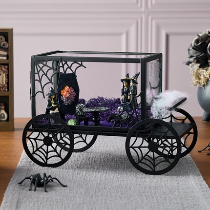 Decorative carriage with black spiderweb-patterned wheels and glass sides, featuring miniature witches, a coffin with orange flowers, purple moss, and a black cat inside.