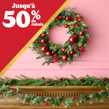 Couronne verte avec ornements rouges sur un mur rose au-dessus d'une cheminée en bois avec une guirlande de sapin, jusqu'à 50 % de réduction en texte blanc sur un triangle rouge.