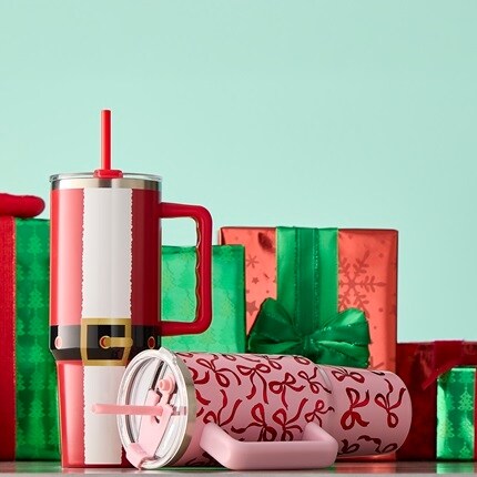 Two tumbler cups: one that is pink and covered in red bows and the other this is a print of Santa's jacket