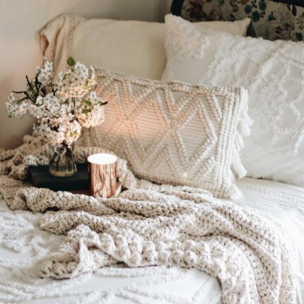 MakerPlace by Michaels Cozy bedroom scene with knitted blankets and textured pillows on a bed. A small vase of white flowers and a lit candle add a serene, warm ambiance.