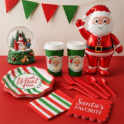 Festive holiday setup with Santa-themed decor, including an inflatable Santa, snow globe, red and green plates and cups, utensils, and party banner.