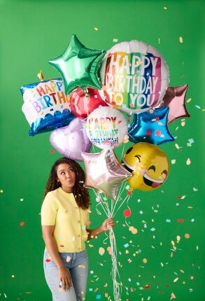woman in yellow shirt holding bouquet of birthday balloons on green background with confetti