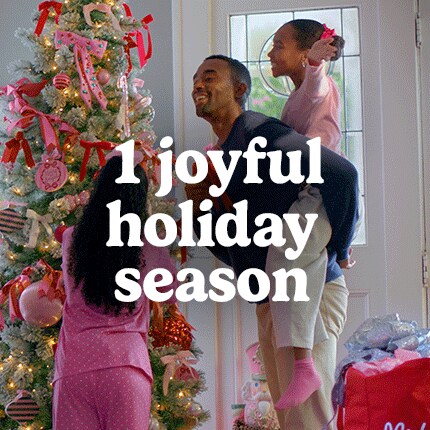 A joyful family decorates a Christmas tree with pink ornaments and ribbons. A man happily lifts a child; warm holiday scene exudes love and togetherness.