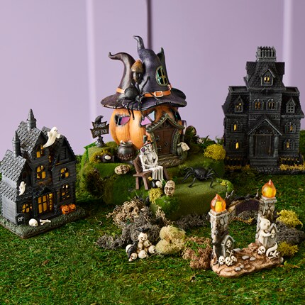 Miniature scene featuring three spooky houses, including a pumpkin-shaped house with a witch hat, surrounded by skeletons, skulls, a spider, and ghost decorations.