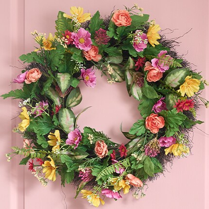 Floral wreath featuring a mix of green leaves and colorful flowers in shades of pink, yellow, orange, and red against a soft pink background.