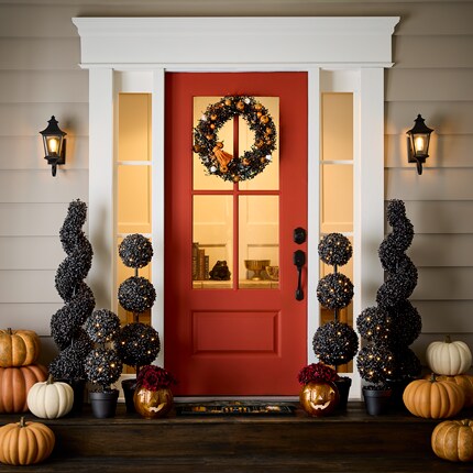 Front porch decorated for Halloween with a red door, black spiral topiary plants adorned with string lights, and a black wreath featuring orange accents. 