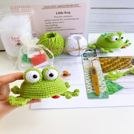 MakerPlace by Michaels A hand holds a crocheted green frog. Behind it is a crochet amigurumi kit with green and white yarn, a crochet hook, and a card detailing the kit's contents. The scene is bright and cheerful, conveying creativity and craft.