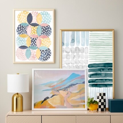 three poster frames with pastel artwork above console with gold lamp