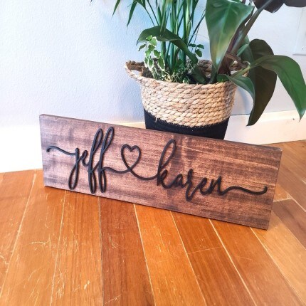 MakerPlace by Michaels Perryhill Rustics A wooden sign with cursive writing reads "jeff ♥ karen" next to a woven basket with a leafy plant. The scene is warm and rustic on a wooden floor.