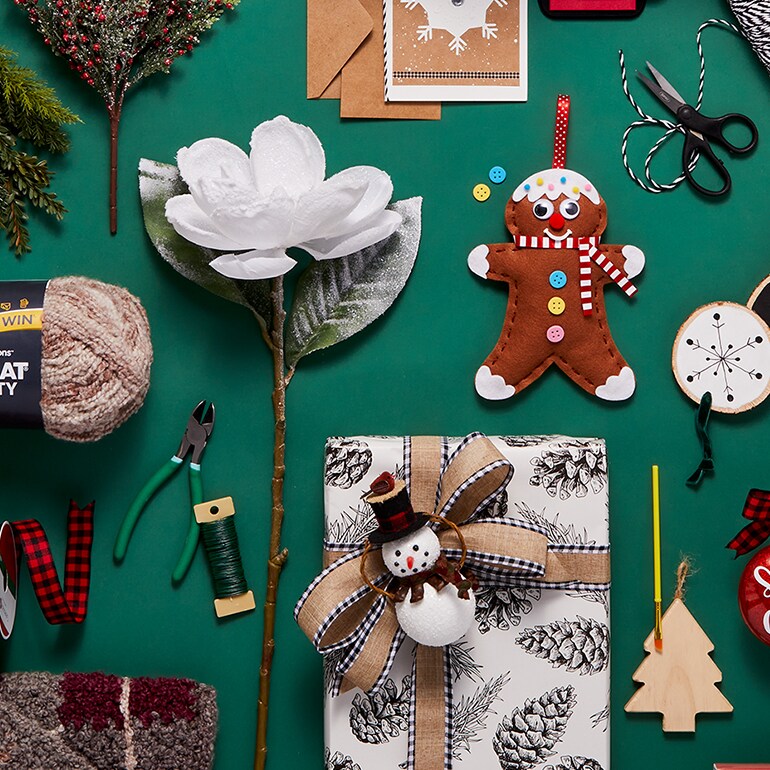 Michaels DIY Festive Christmas craft flat lay on green: a red “Santa Cam” ornament, baubles, gingerbread figure, white flower, fabric, and sewing tools.