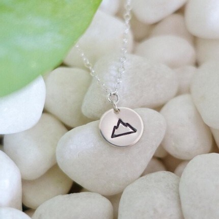 MakerPlace by Michaels Delicate silver necklace with a mountain pendant rests on smooth white pebbles beside a green leaf, creating a calm, natural mood.
