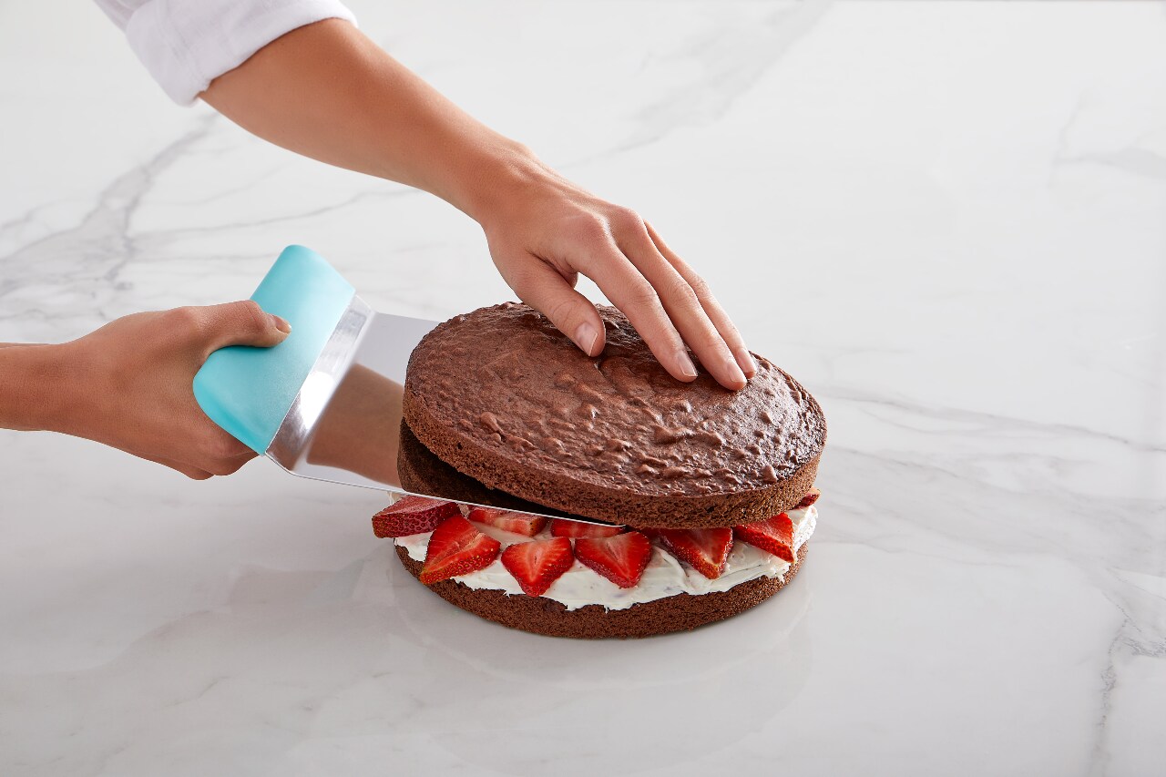 A pair of hands places a layer of cake onto a stack of strawberries and cream