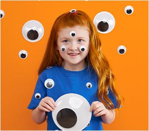 Little girl with googly eyes on her face, holding a large googly eye against an orange background