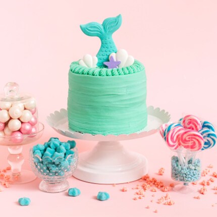 teal mermaid themed cake on stand surrounded by candy with pink background