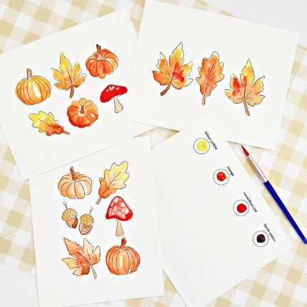 MakerPlace by Michaels Autumn watercolor cards with pumpkins, maple leaves and mushrooms surround paint swatches and brushes on a checkered tablecloth.
