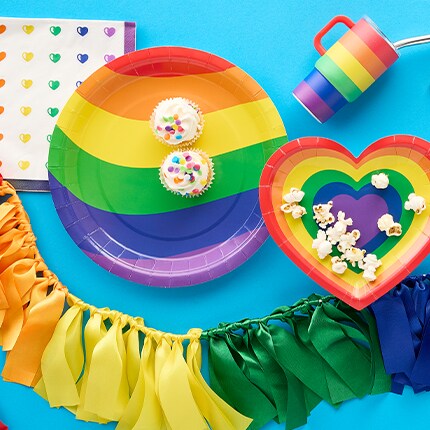 Rainbow-striped paper plates, a heart-shaped plate with popcorn, a rainbow-striped cup, and a napkin with multicolored heart patterns on a bright blue background.