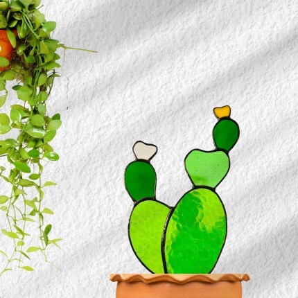 MakerPlace by Michaels Cheerful cartoon cactus with green pads and tiny buds in a terracotta pot, beside a trailing houseplant against a white textured wall.